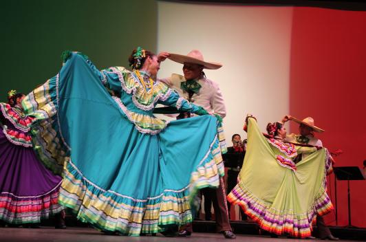Couples dancing in front of a projection of the Mexican flag. Some a wearing large skirts that they are holding wide, while others are in hats