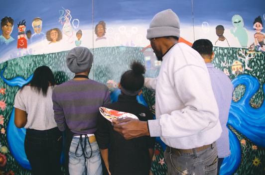 Various people working on a mural depicting a valley with happy faces and green grass