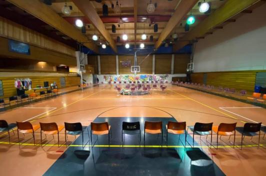In a gymnasium, chairs are set out along all four walls. A rack of clothes sits behind one set of chairs.
