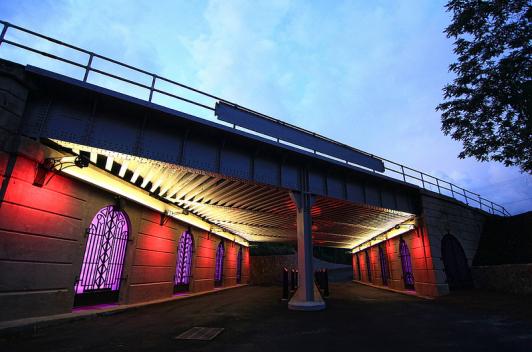 A bridge at dusk. Under the bridge, along the walls are iron gates which are lit up from behind in purple. 