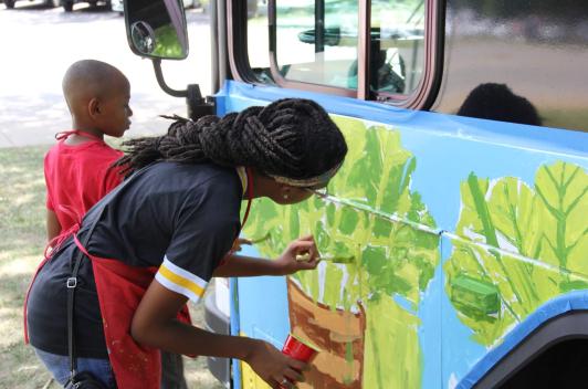 Two young people paint vegetables on the side of a bus.