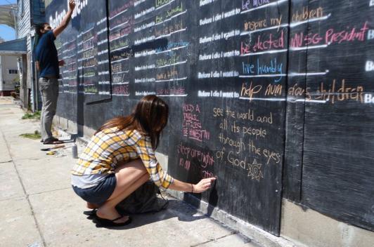 At a chalk wall with prompts one person stands and another sits writing on the wall.