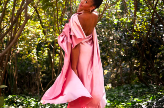 A person in a pink, silk dress, stands with their back to the camera, turning only their face and smiling. They are standing in a large forest.