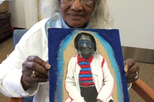 A person sits smiling, holding a painting of themselves.