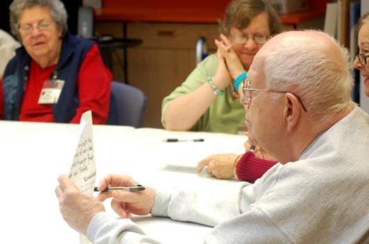 A number of older people sit around a table, reading from papers.