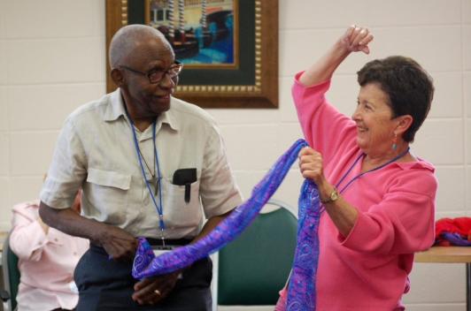 Two older people stand, smiling, holding on to a blue streamer.