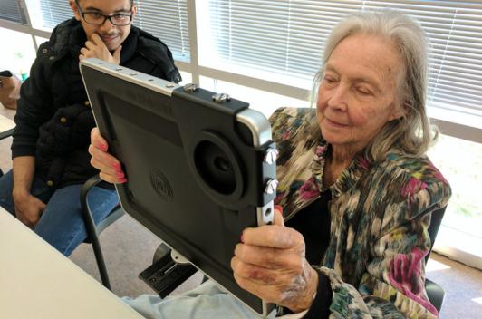 An older person sits by a young person. The older person is holding up a tablet and watching something on it.