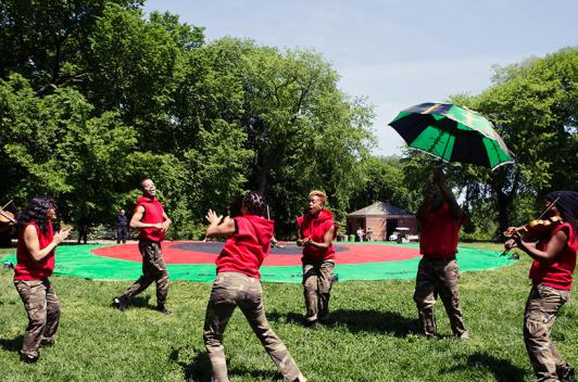People stand in a circle in a red shirts and khaki in a park. Some are dancing, some are playing instruments.