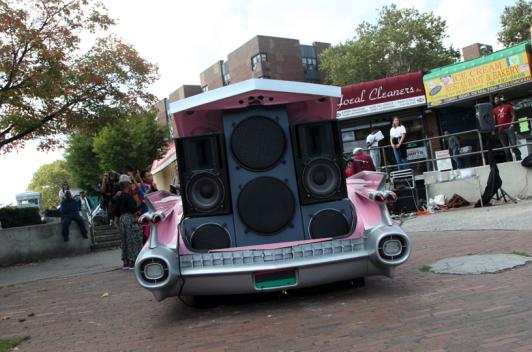 The trunk of a pink car is open and has four or five different speakers piled on top of each other.