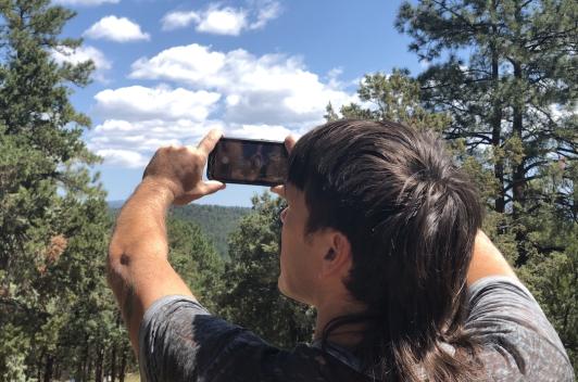 A person stands in the forest, holding a phone taking a picture.