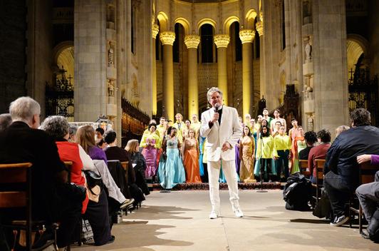 In a church, a person in a white suit stands at the front. Behind are people in colorful clothing and in the front are people sitting in the pews.