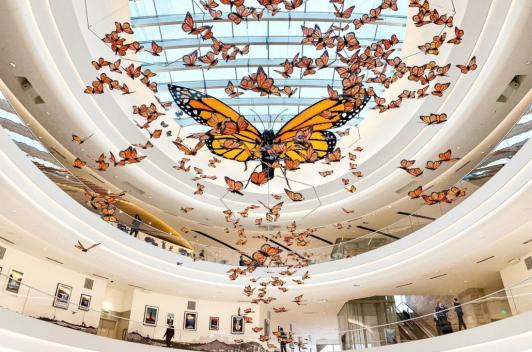 In a large, open atrium with a sky light, a display of monarch butterflies in various sizes hang from the ceiling.