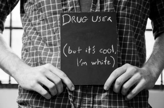 Without showing the face, a person stands holding a sign say, Drug User (but it's ok I'm white).