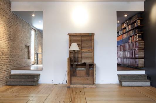 Facing a white wall, there are two hallways you can take. One is to an open and sunny space, the other is to a library like space with lots of books. In the middle of the wall is an old shoe sign seat.