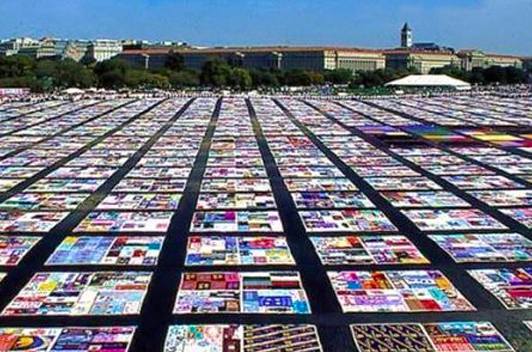 In an open field by a low level building, are many colorful quilts laid out.