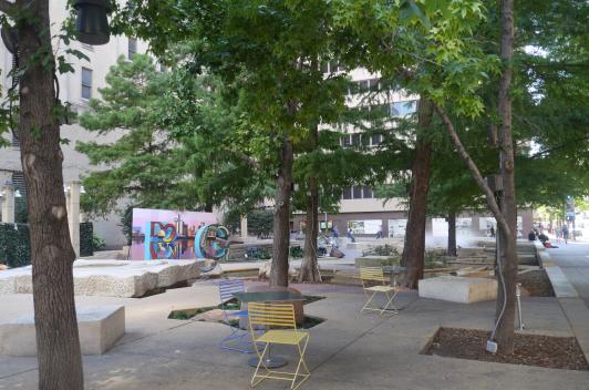 A relaxing concrete park in the middle of the city. The park has tables and chairs along with some grass and trees, water features, and sculptures.