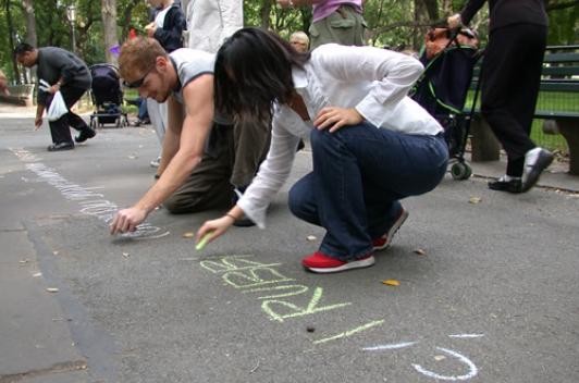 Two people lean over to write words in chalk on the pavement.