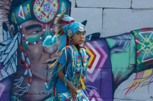 A child in traditional wear stands in front of a mural depicting a person in similar traditional wear.