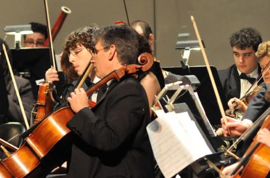 The strings section of an orchestra sits ready to play. The orchestra is made up of both adults and children.