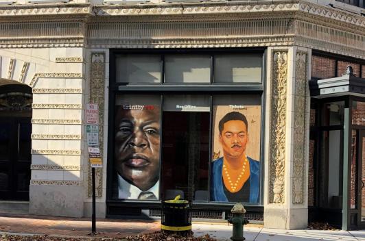 In the window of a building, facing the street, sits two portraits of two different people.