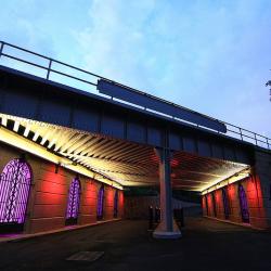 A bridge at dusk. Under the bridge, along the walls are iron gates which are lit up from behind in purple. 