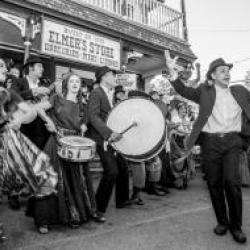 A black and white photo features a group of people dancing and playing instruments in front of a store.