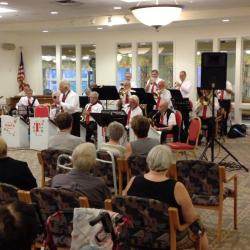 A brass band in white collared shirts and red ties sits and plays at the front of a room. Another group of people sit around the room and enjoy the music.