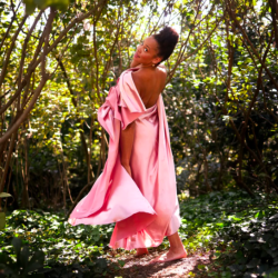 A person in a pink, silk dress, stands with their back to the camera, turning only their face and smiling. They are standing in a large forest.