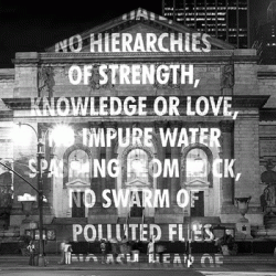 A black and white photo shows an older building with arches and columns with words projected on the front of it.