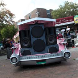 The trunk of a pink car is open and has four or five different speakers piled on top of each other.