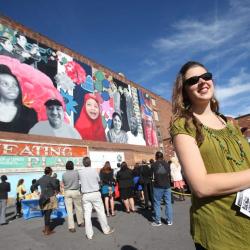 Person smiling infront of a large mural on a brick building in the background. People are gathered behind them, facing the mural.