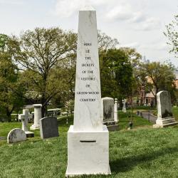 In a graveyard with green grass is a large marble obelisk.