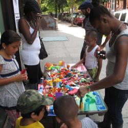 In front of a laundromat, people are gathered around a table filled with arts and crafts items.