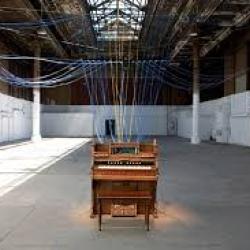 In an empty warehouse sits a brown piano. Strings go from the piano to the ceiling.