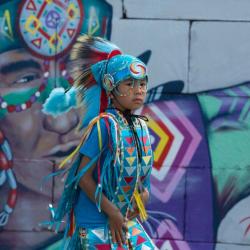 A child in traditional wear stands in front of a mural depicting a person in similar traditional wear.