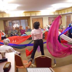 Person standing in the middle of a room holding many colored scarfs, which others who are sitting in a circle grab a hold of