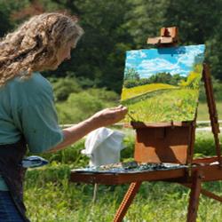 In a green pasture, a person stands at a table that holds an easel and is painting the landscape.
