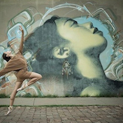 Mural of a person's face looking towards the sky. Beside the mural a person does a ballet move, looking towards the sky in a similar fashion to the mural.