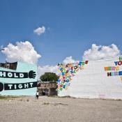 Two buildings beside each other. Both have murals painted on them. One has a light blue background and the text hold tight written on to arms. The other has a white background and magnetic colorful letters.