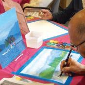 An older person sits at a table with a red table cloth. They are painting a landscape.