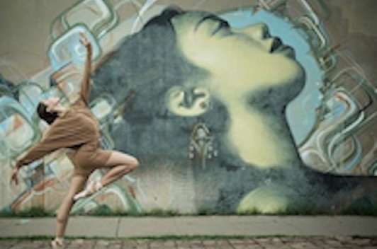 Mural of a person's face looking towards the sky. Beside the mural a person does a ballet move, looking towards the sky in a similar fashion to the mural.
