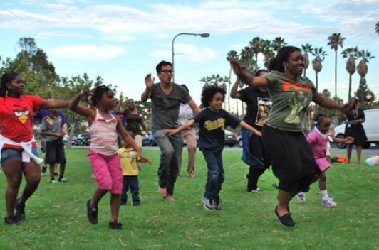 A group of adults and children dance on a green lawn.