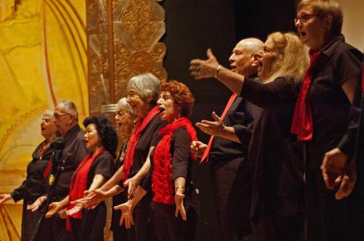 A group of older people standing on a stage in black outfits with red scarfs, singing