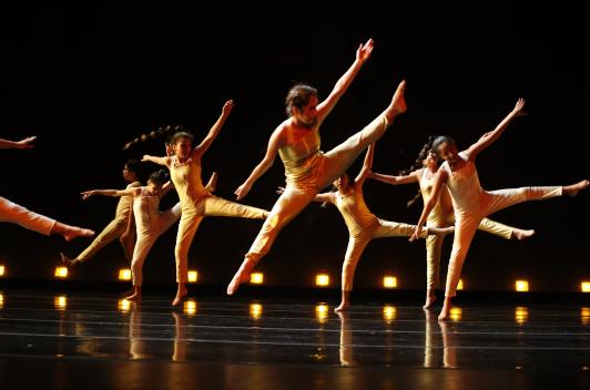 Students in gold outfits dance on a low lit stage