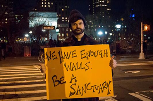 A person standing near a crosswalk holding a sign that says, "We have enough walls. Be a sanctuary."