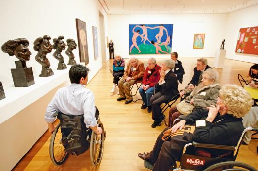 Photo of a person in a wheel chair guiding others through an art exhibit