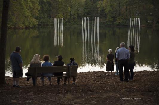 People standing and sitting by a lake that has thin poles emanating light coming up from the water.