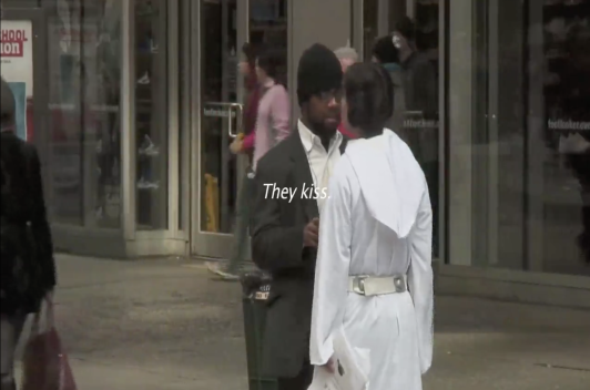 A person in a suit and black beanie and a person in a white Princess Leia costume stand in front of each other on a street. Text reads, they kiss.