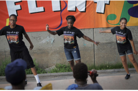 Three dancers stand on a platform, dancing in sync. Behind them is a colorful banner with the word, Flint, on it.