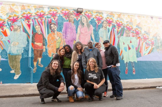 A small group gathered in front of a mural posing and smiling.